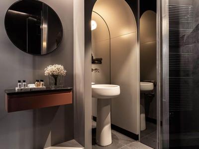 Modernes Badezimmer mit rundem Spiegel, Waschbecken und dekorativem Blumenstrauß auf einem Wandregal.