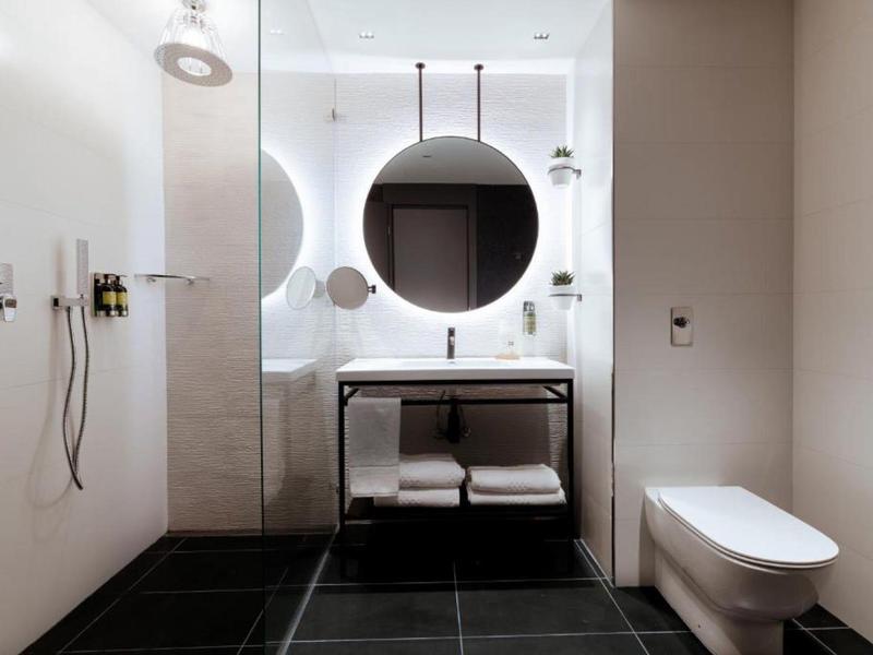 Modern hotel bathroom with round backlit mirror, glass shower, and wall-mounted toilet.