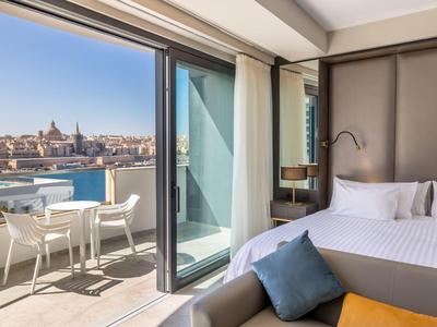 Chambre d'hôtel moderne avec terrasse et vue sur une ville et l'eau par ciel clair.