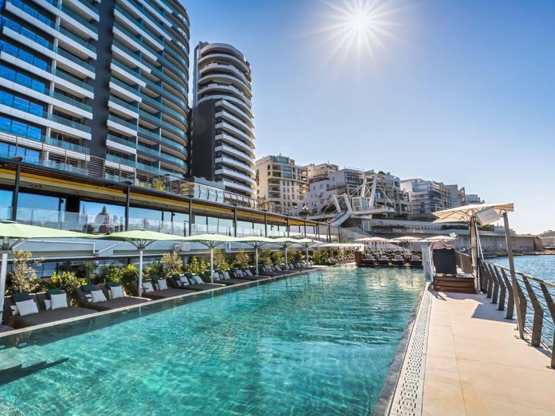 Piscine d'hôtel moderne avec transats à côté d'une piscine claire et vue sur la mer au soleil.