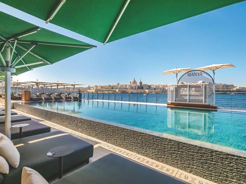 Piscine à débordement avec coin salon donnant sur une ville au bord de l'eau sous un ciel clair.