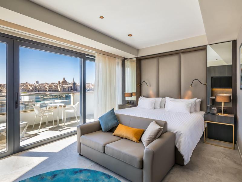 Chambre d'hôtel moderne avec lit double, canapé et fenêtres du sol au plafond donnant sur une terrasse avec vue sur la ville.