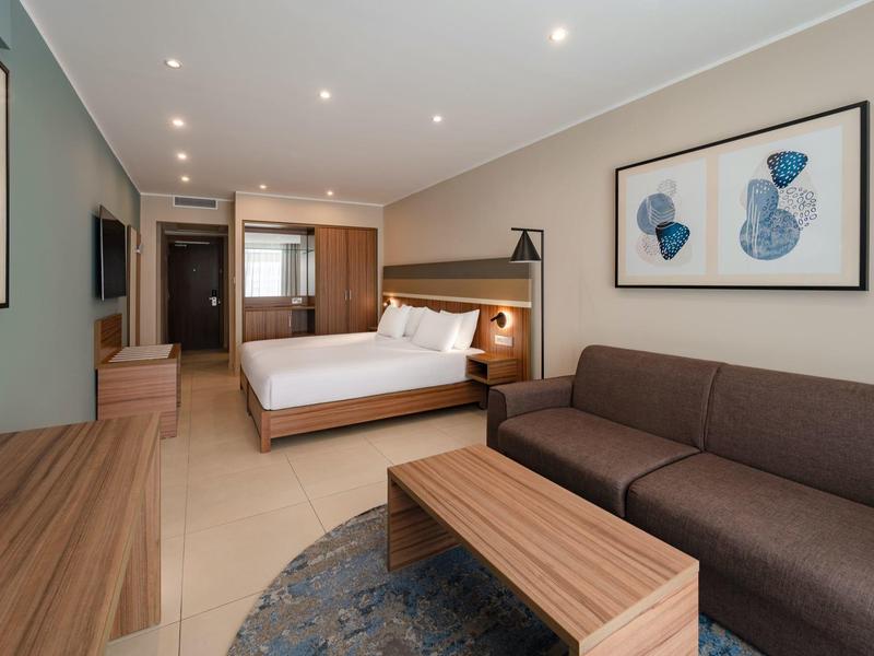 Habitación de hotel moderna con cama, sofá y muebles de madera en tonos neutros.