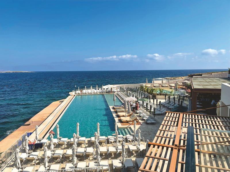 Piscina infinity con tumbonas junto al mar bajo un cielo azul despejado.