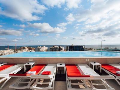 Piscina a sfioro su terrazza panoramica con lettini rossi e vista sulla città e sul mare.