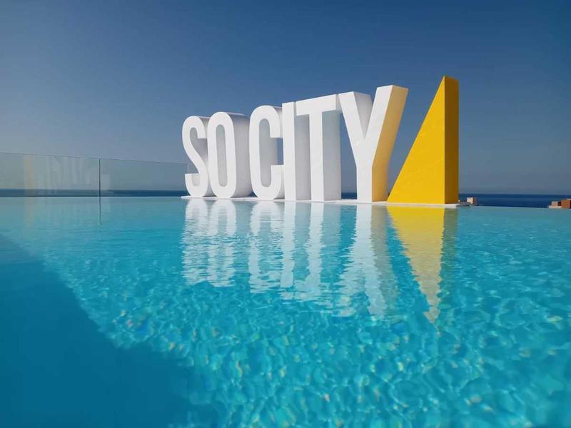 Grande insegna bianca e gialla So City ai bordi di una piscina limpida sotto un cielo blu.