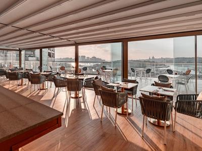 Restaurant mit Glasfront, Holztischen, Korbsesseln und Blick auf Hafen mit Booten bei Sonnenuntergang.