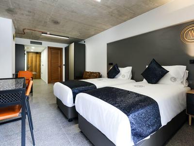 Nowoczesny pokój hotelowy z dwoma łóżkami, biurkiem i betonowym sufitem.