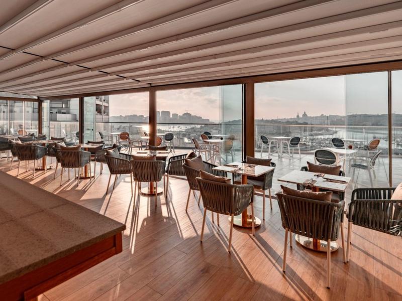 Nowoczesna restauracja z panoramicznym widokiem na port i jachty o zachodzie słońca.