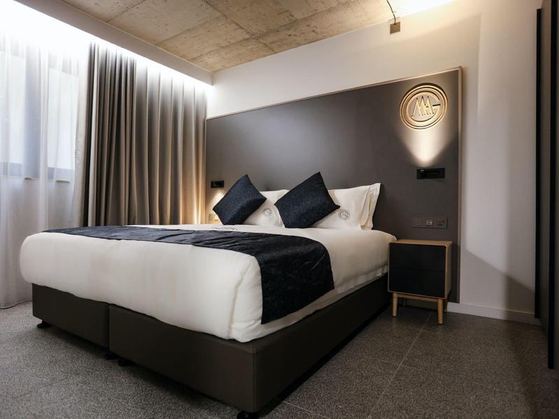 Nowoczesny pokój hotelowy z dużym łóżkiem, białymi i ciemnymi poduszkami oraz prostym stolikiem nocnym.