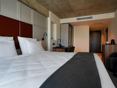 Modern hotel room with large bed, white linens, wooden furniture, and minimalist decor.
