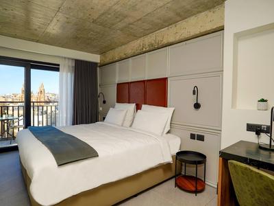 Modern hotel room with large bed, balcony, and city view under a concrete ceiling.