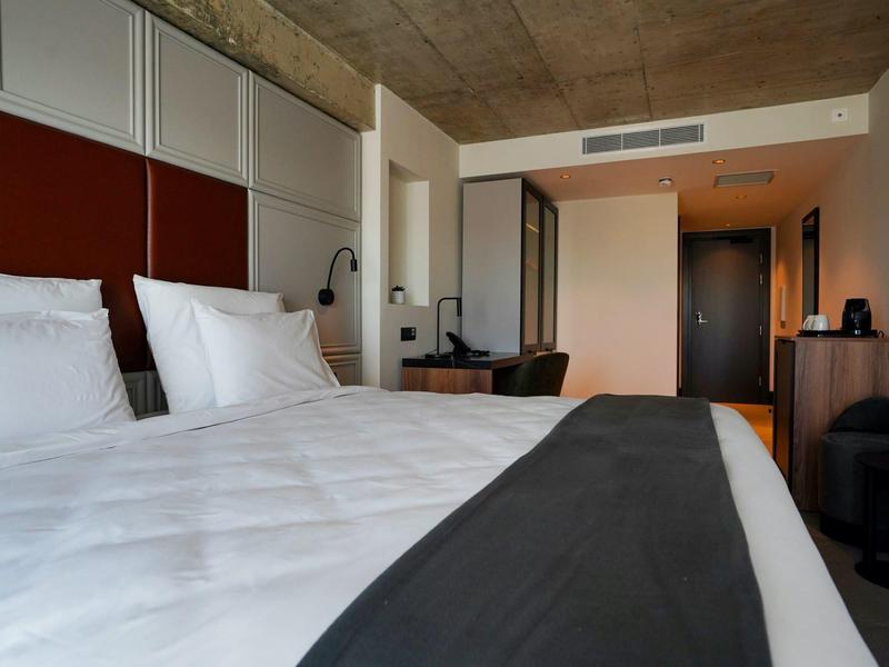 Modern hotel room with large bed, white linens, wooden furniture, and minimalist decor.