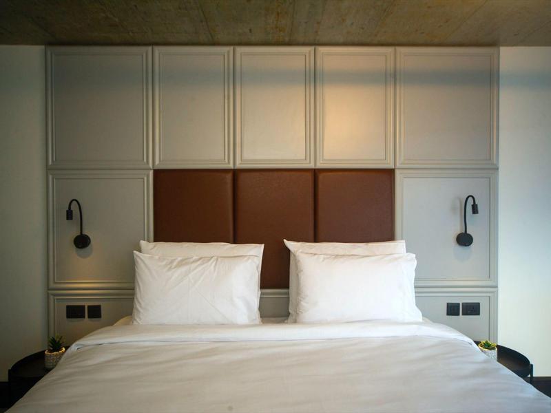 Minimalist hotel bedroom with white bedding, brown padded headboard, and wall-mounted reading lights.