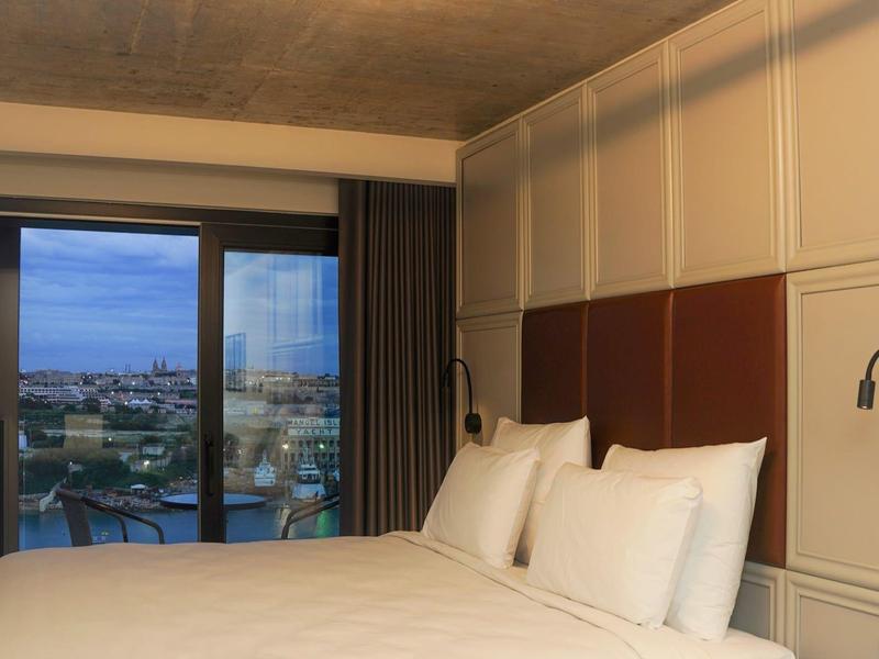 Modern hotel room with large bed, bedside lamps, and city view through sliding glass door.
