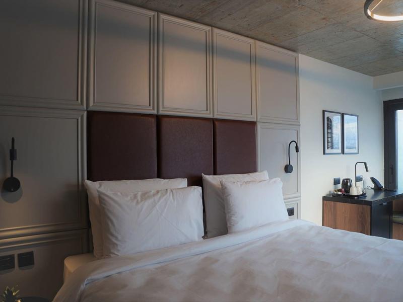 Modern hotel room with double bed, pillows, bedside lamps, and a small desk near a balcony.