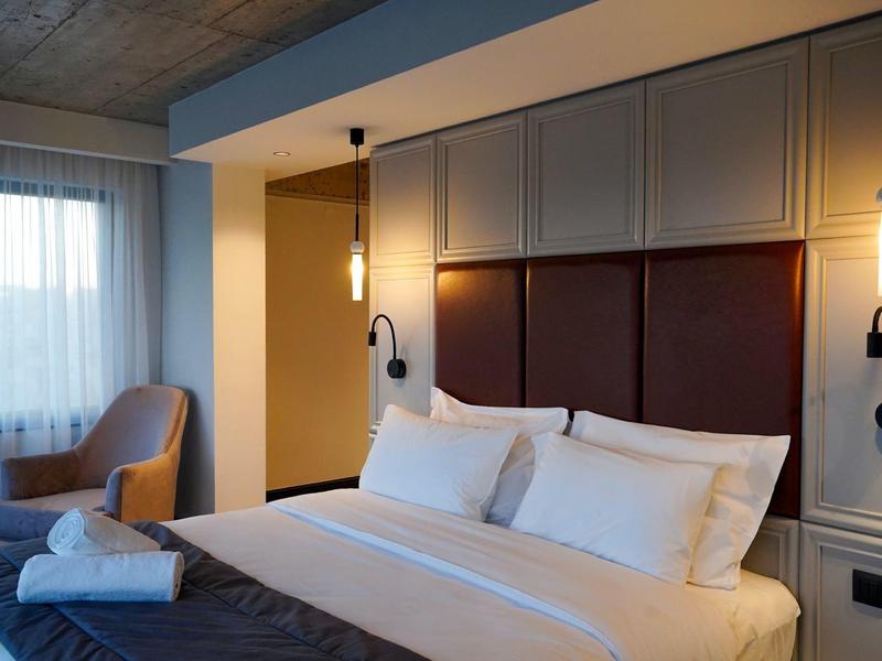 Hotel room with large bed, soft lighting, cozy chair, and window with sheer curtains.