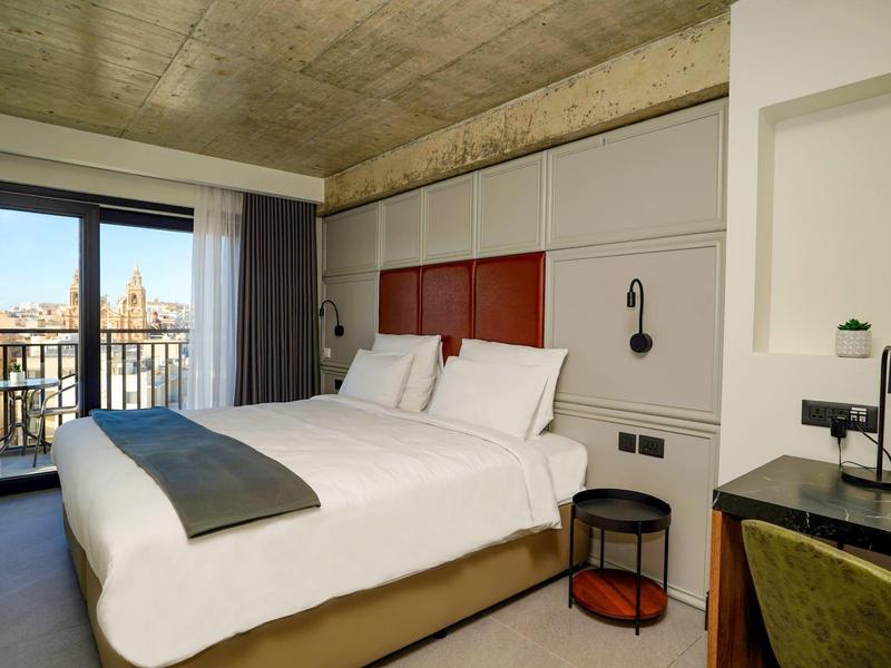 Modern hotel room with large bed, balcony, and city view under a concrete ceiling.