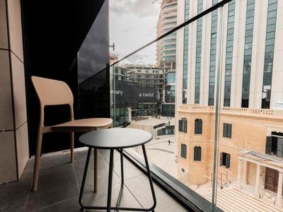 Balkon mit Holzstuhl und kleinem runden Tisch vor Glasgeländer, im Hintergrund Hochhäuser.
