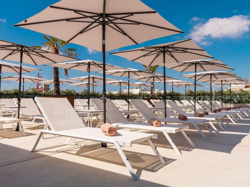 Mehrere weiße Liegestühle und große Sonnenschirme stehen in einer Reihe unter blauem Himmel.