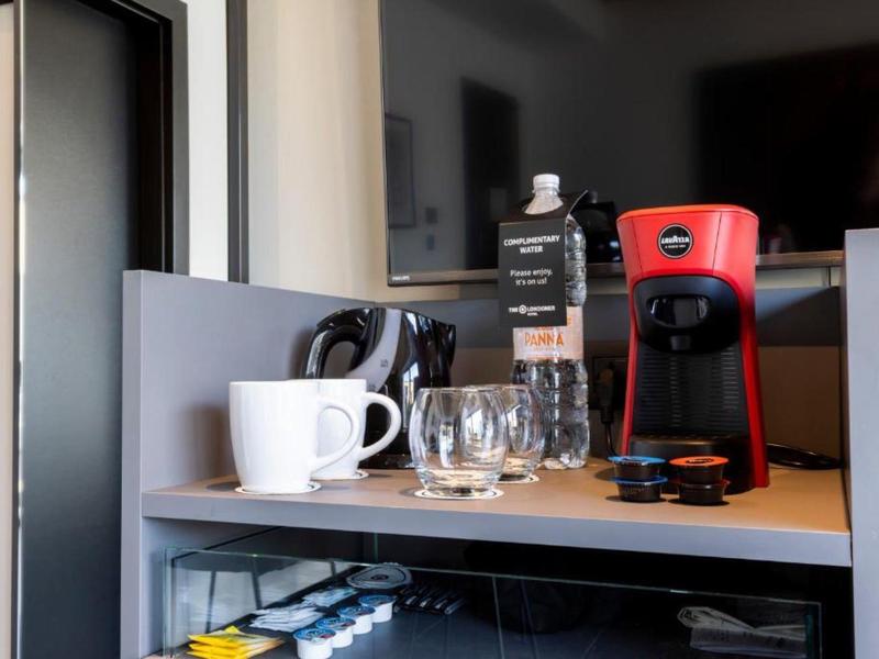 Ein roter Kaffeautomat, weiße Tasse, Teekanne und Gläser stehen auf einem grauen Regal vor einem Fernseher.