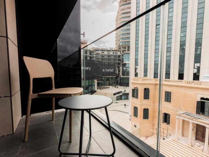 Balkon mit Holzstuhl und kleinem runden Tisch vor Glasgeländer, im Hintergrund Hochhäuser.