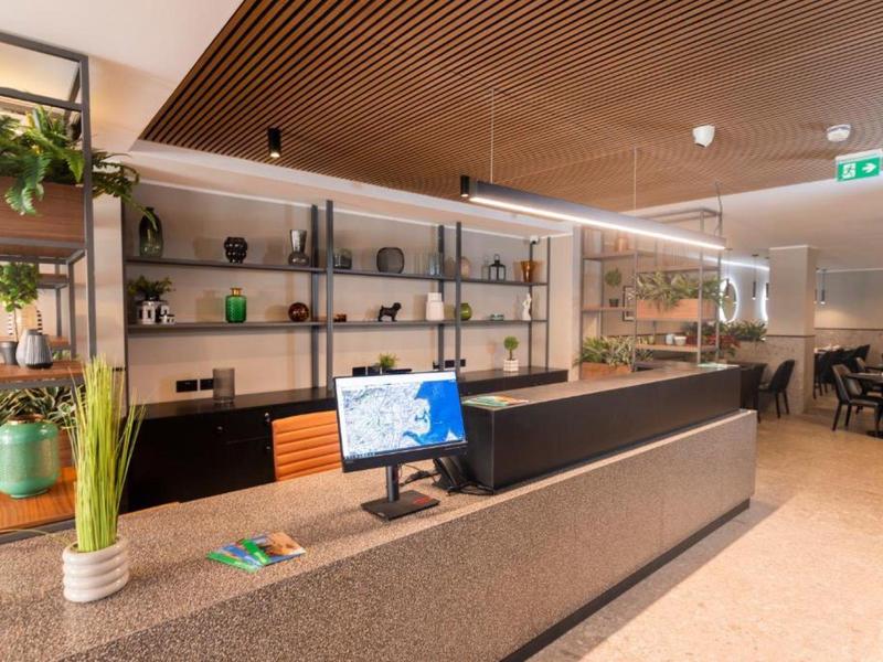 Moderner Empfangsbereich mit Theke, Computer, Regalen und grünen Pflanzen in hellem Raum.