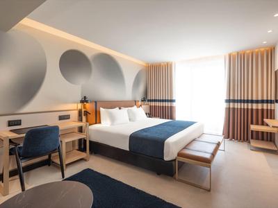 Modernes Hotelzimmer mit großem Bett, Schreibtisch, Sessel und großem Fenster mit Vorhängen.