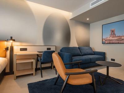 Modern eingerichtetes Hotelzimmer mit Sitzbereich, Schreibtisch und abstrakter Wandgestaltung.