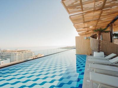 Infinity-Pool mit Liegestühlen und Blick auf Stadt und Meer unter klarem Himmel