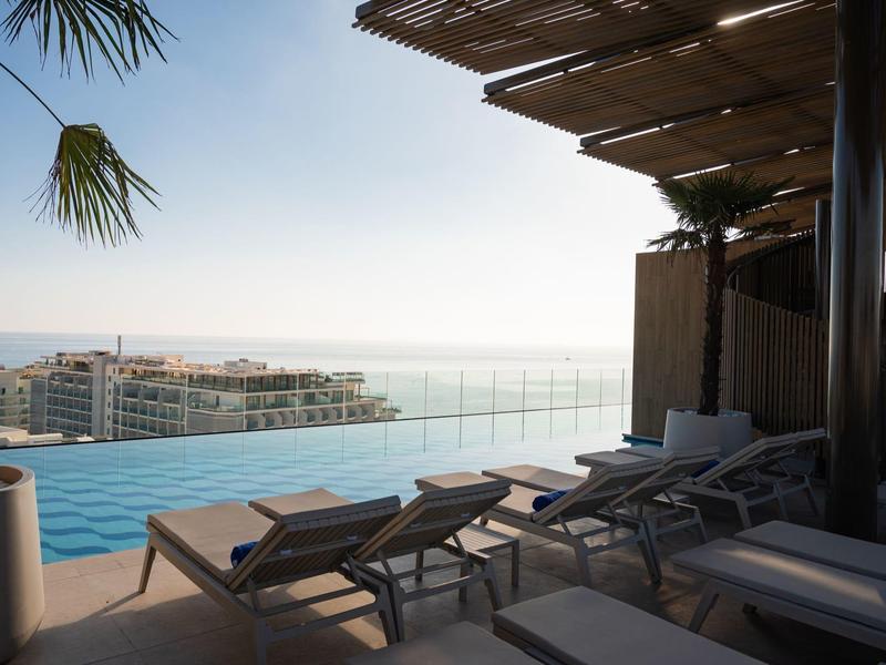 Infinity-Pool mit Liegestühlen und Blick auf die Stadt und das Meer am Horizont.