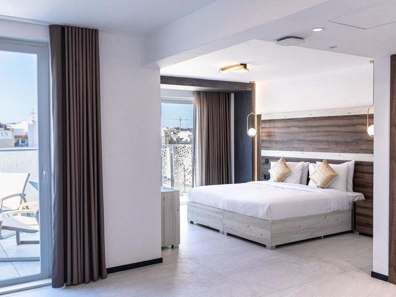 Bright hotel room with large bed, wooden headboard, and balcony with outdoor chairs.