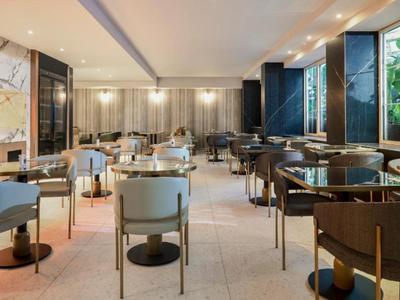 Área de restaurante de hotel decorada modernamente con mesas, sillas y grandes ventanas con vista al verde.