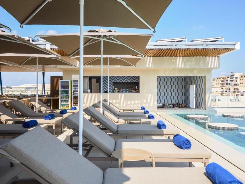 Área de piscina moderna con tumbonas, toallas azules y grandes sombrillas en un hotel.