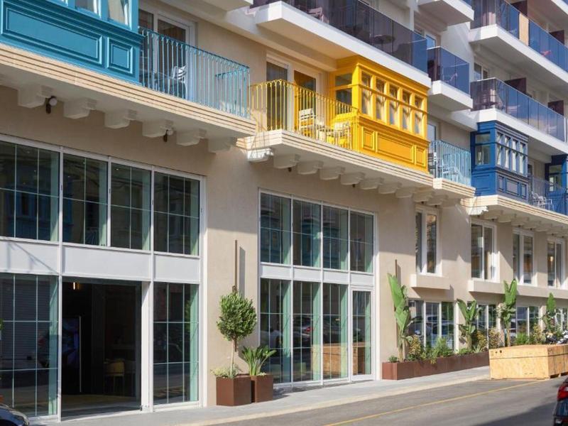 Fachada del hotel con grandes ventanas, balcones y barandillas coloridas en amarillo y azul.