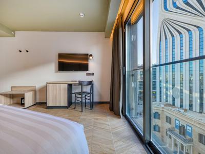 Hotelzimmer mit Bett, Schreibtisch, Stuhl und großem Fenster mit Stadtblick.