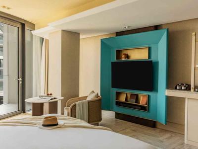 Moderní hotelový pokoj s velkým oknem, televizorem v tyrkysovém rámu a pohodlným nábytkem.