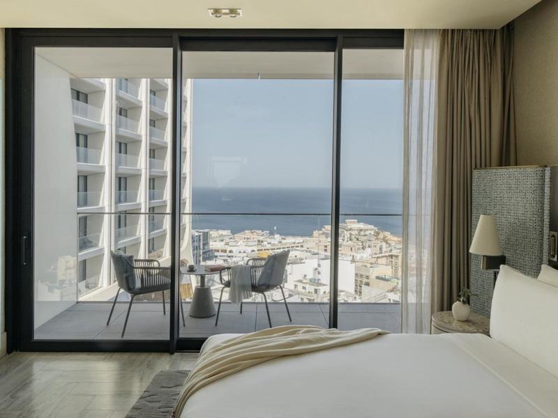 Ložnice s dvojlůžkem a výhledem na balkon, město a moře přes velká skleněná okna.