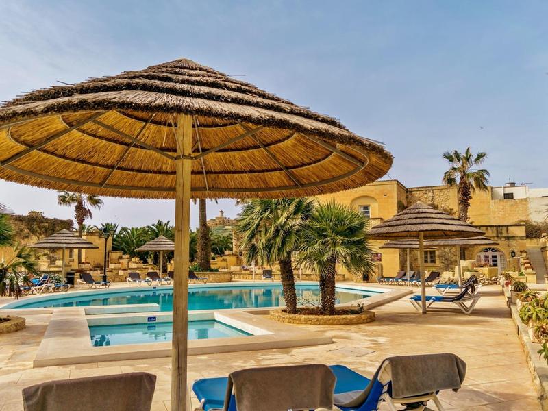 Bazénová zóna s slunečníky ze slámy, lehátky a palmami pod jasnou oblohou.