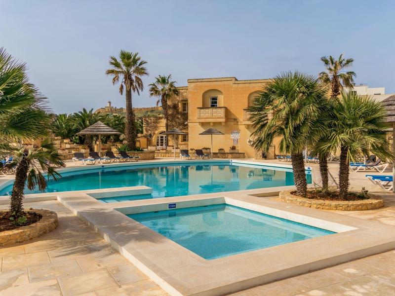 Bazénová oblast se dvěma bazény, palmami a žlutou hotelovou budovou v pozadí.