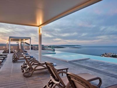 Nowoczesny basen infinity z leżakami na drewnianym tarasie z widokiem na morze o zachodzie słońca.