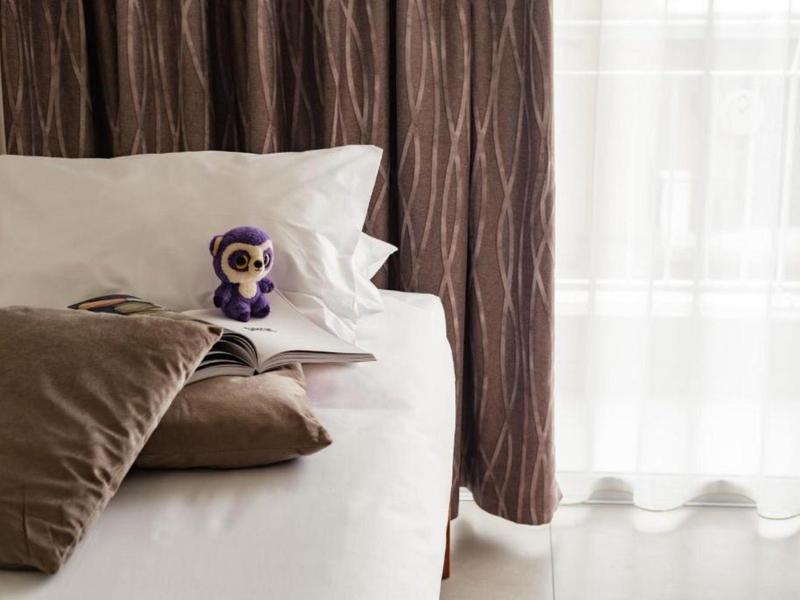 Wit bed met bruine kussens, een klein teddybeertje en doorschijnende gordijnen die licht binnenlaten.