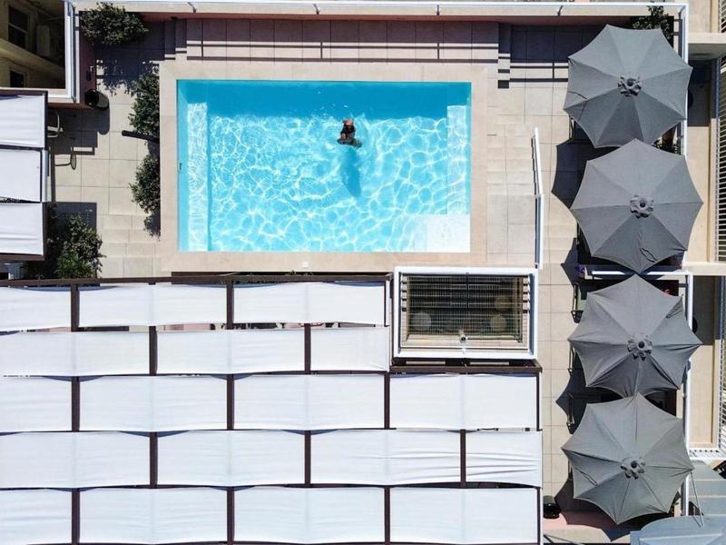 Rechteckiger Pool mit blauen Wasser, Person schwimmt, daneben graue Sonnenschirme und weiße Liegen.
