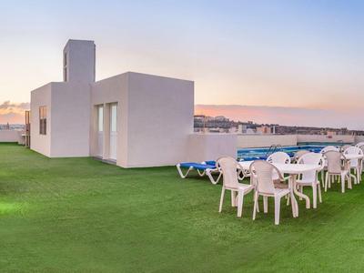 Terrasse sur le toit avec gazon artificiel, chaises et tables blanches offrant une vue sur la ville au coucher du soleil.