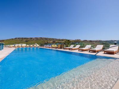 Großer Pool mit Liegestühlen und sonnigem Himmel in einer Ferienanlage mit Hügeln im Hintergrund.