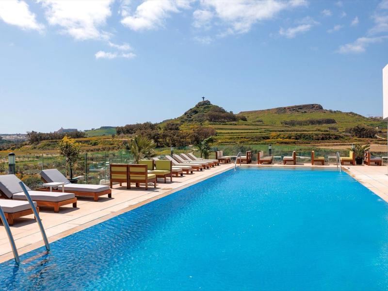 Modern buitenzwembad met ligstoelen en uitzicht op groene heuvels onder een blauwe lucht.