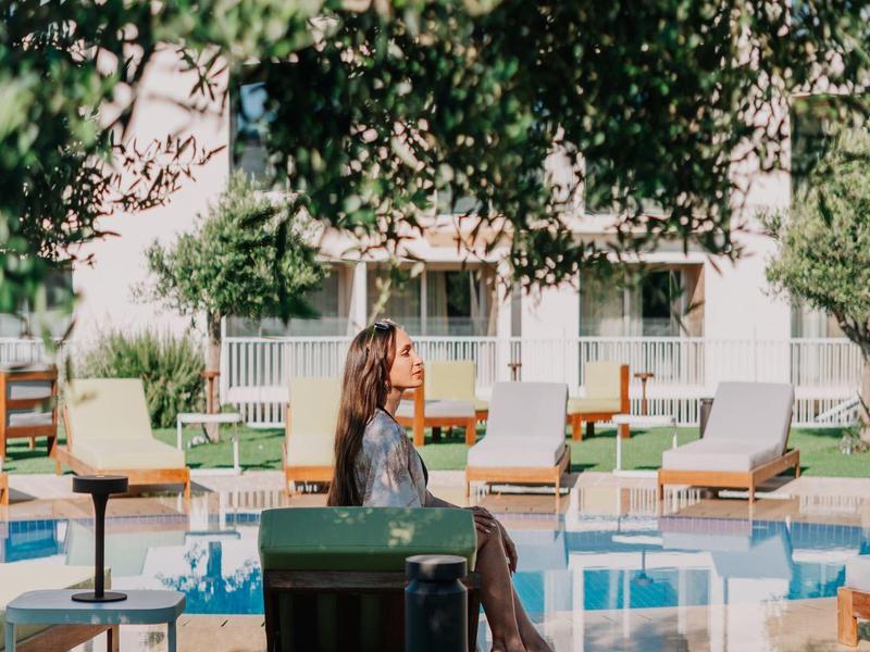 Vrouw zit ontspannen bij het zwembad van een modern hotel met ligstoelen en bomen op de achtergrond.