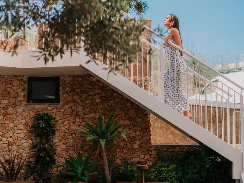 Vrouw in witte jurk loopt een buitentrap op naast een met planten versierd gebouw onder een heldere hemel.