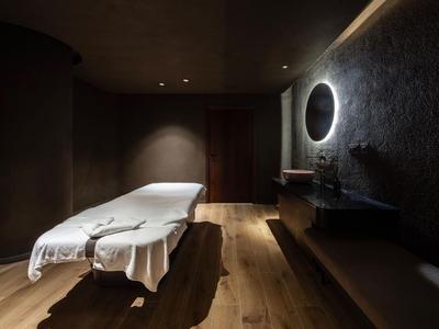Dunkler Wellnessraum mit Massageliege, Holzfußboden und rundem beleuchtetem Spiegel.