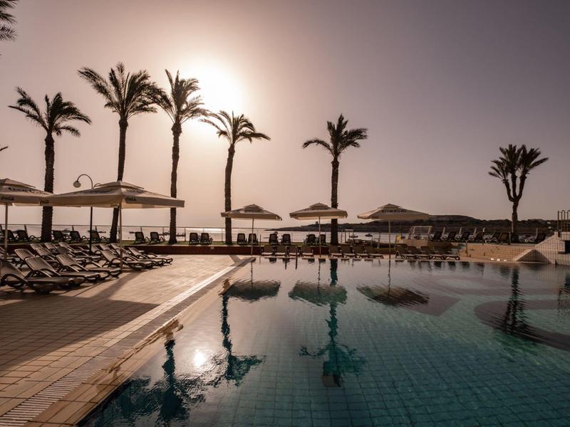 Piscine avec chaises longues et palmiers au coucher du soleil dans un complexe hôtelier.